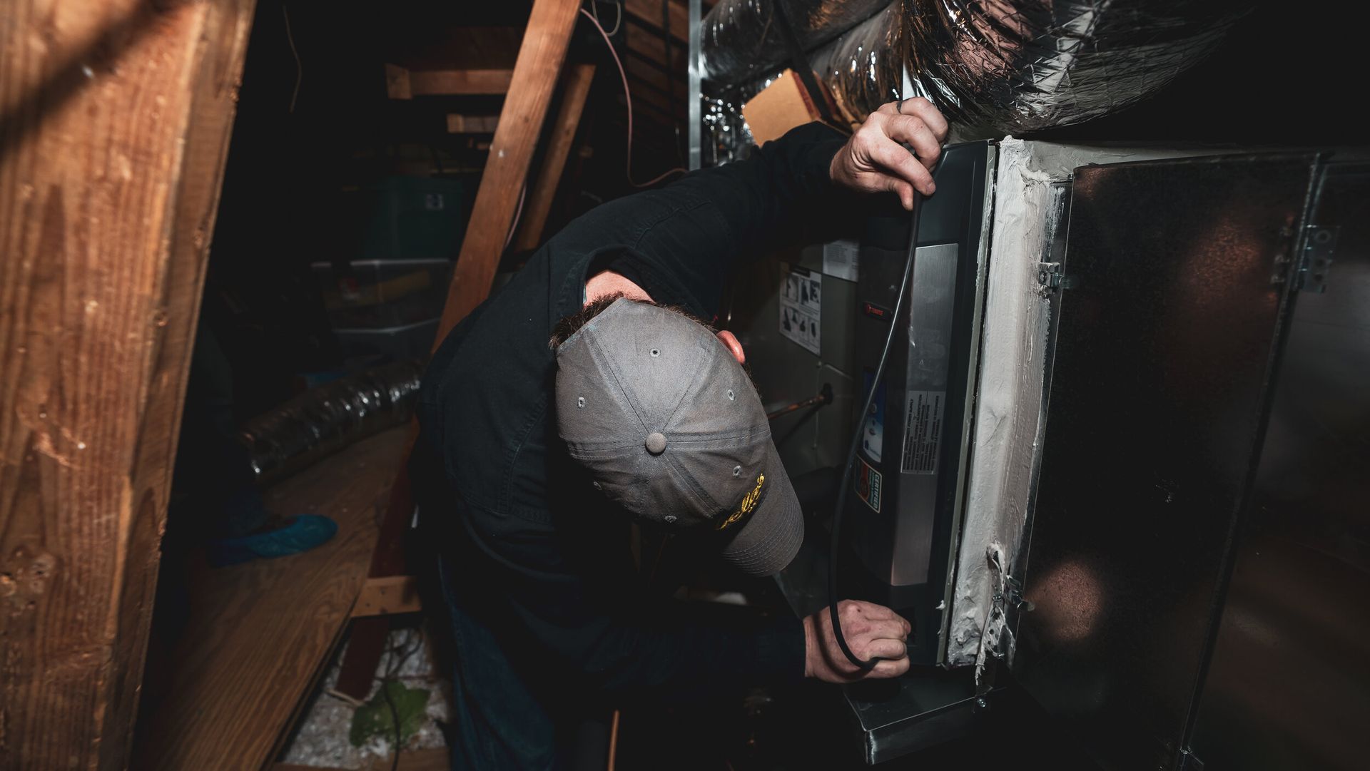 A man in a hat is working on an air conditioner in an attic.