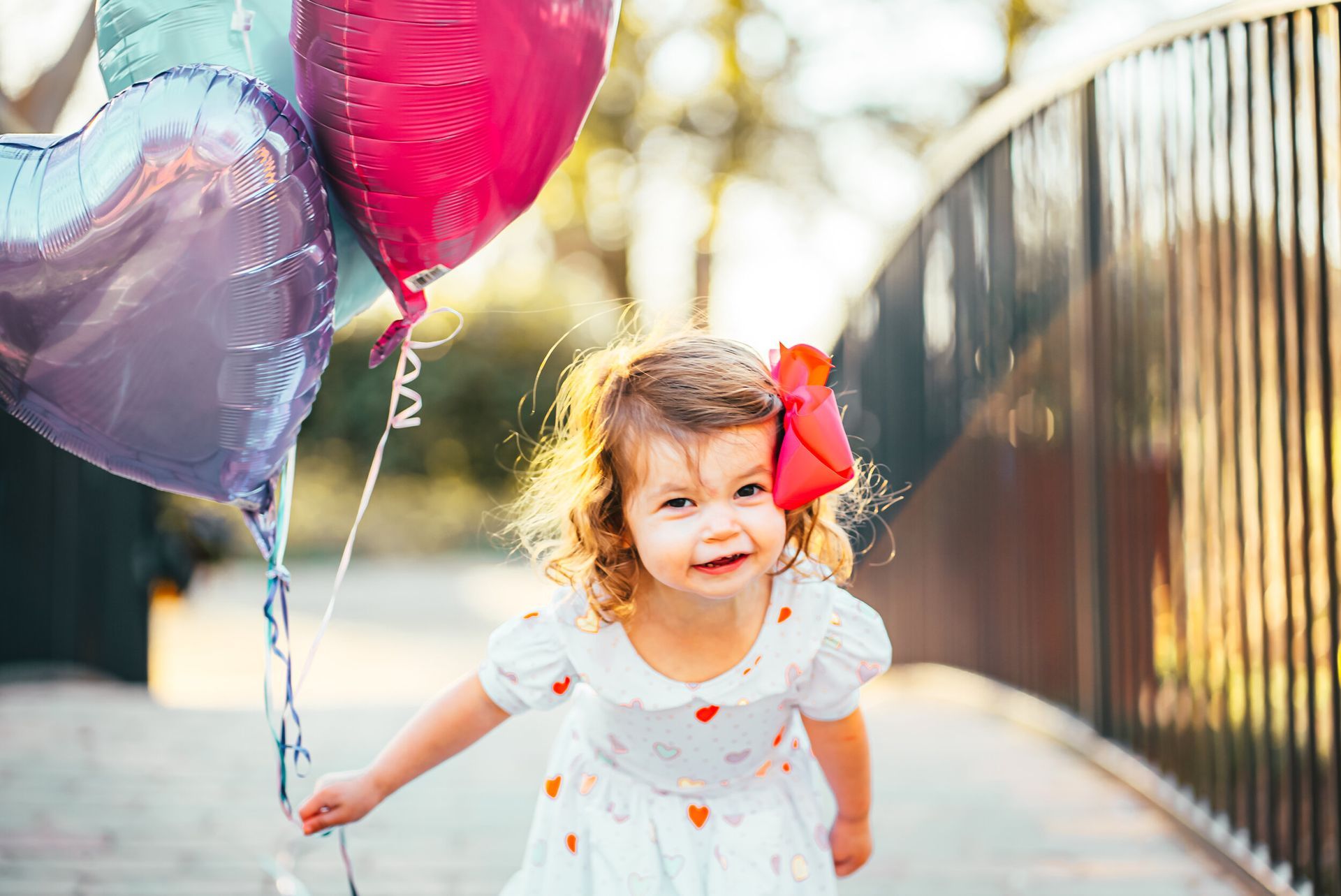 A little girl in a white dress is holding three balloons.