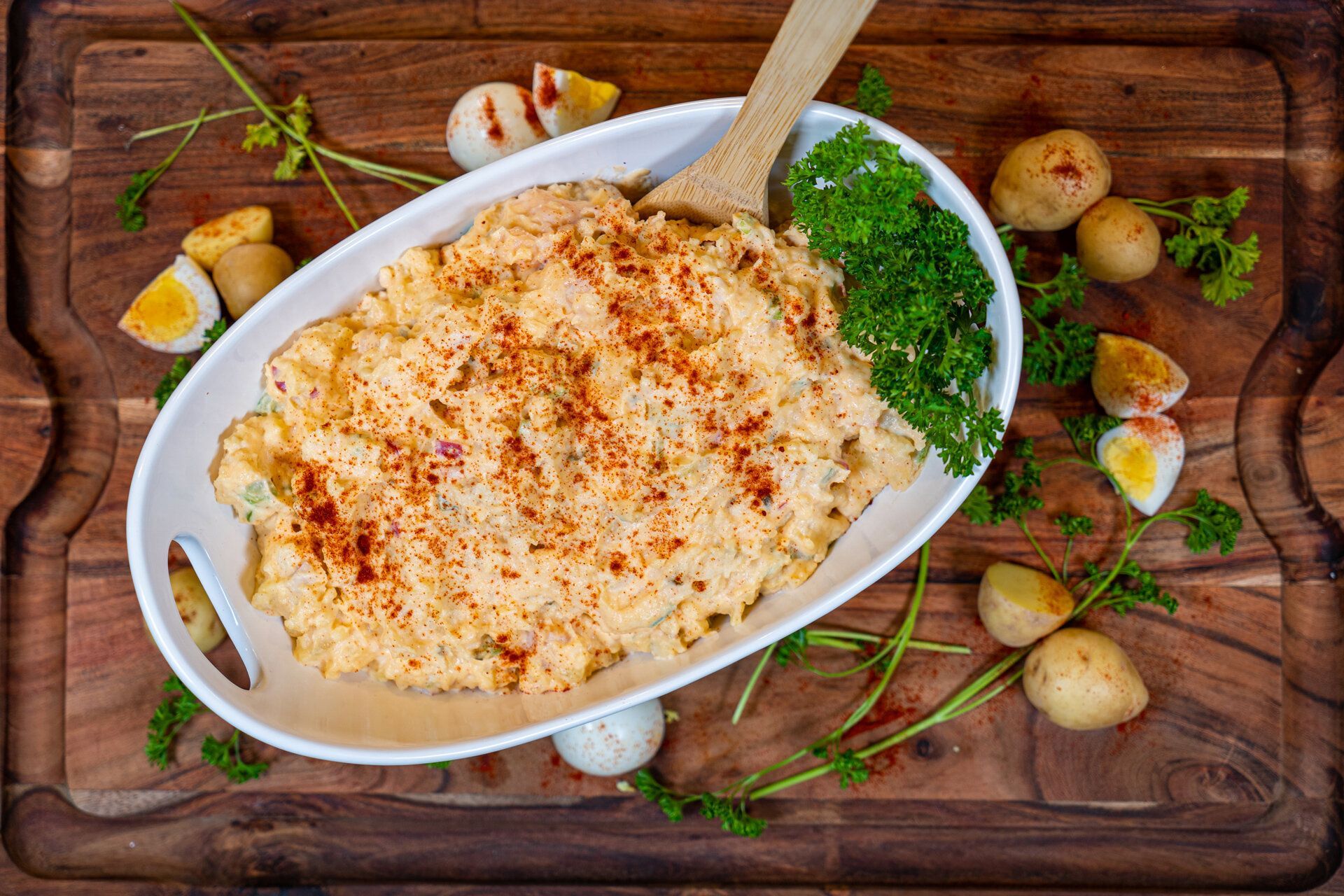 A bowl of potato salad with eggs and parsley on a wooden cutting board.