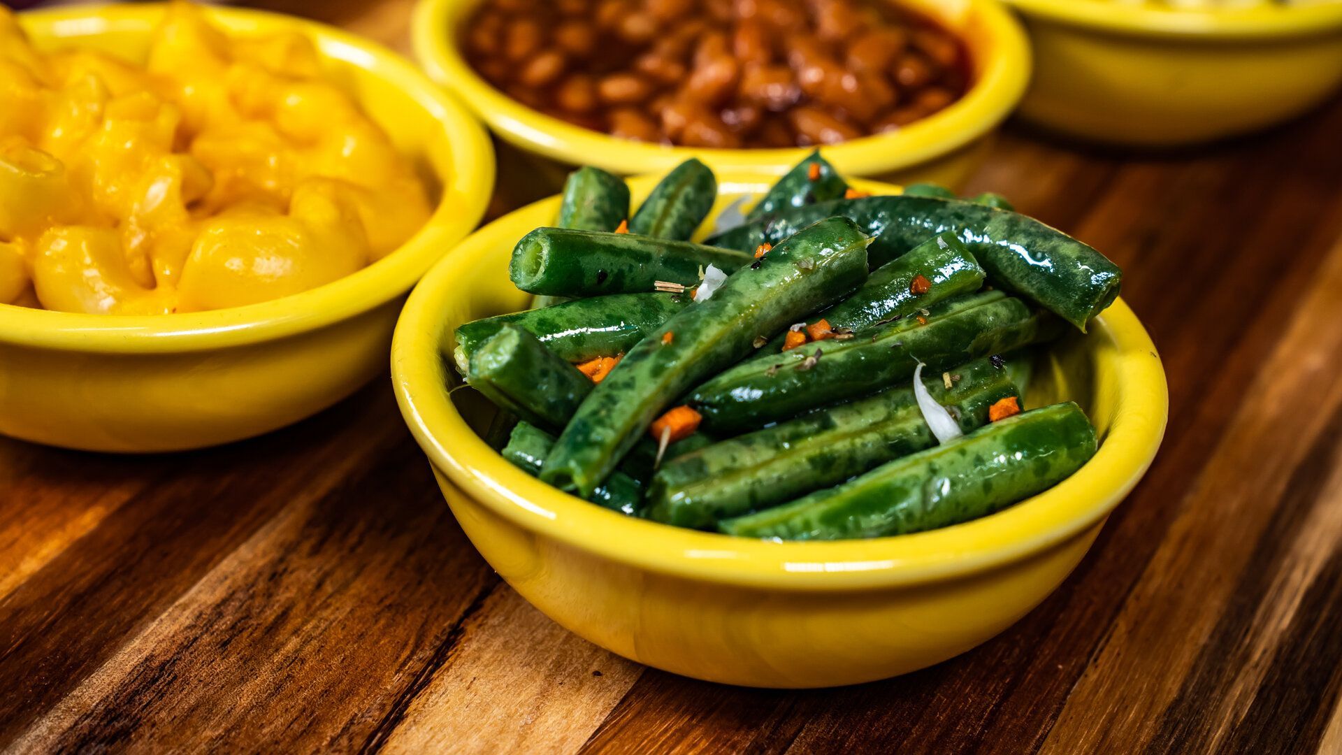 A bowl of green beans is sitting on a wooden table next to other bowls of food.