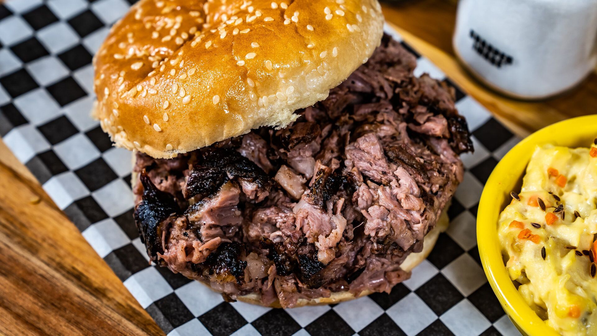 A sandwich with meat on a bun is sitting on a checkered napkin next to a bowl of mashed potatoes.