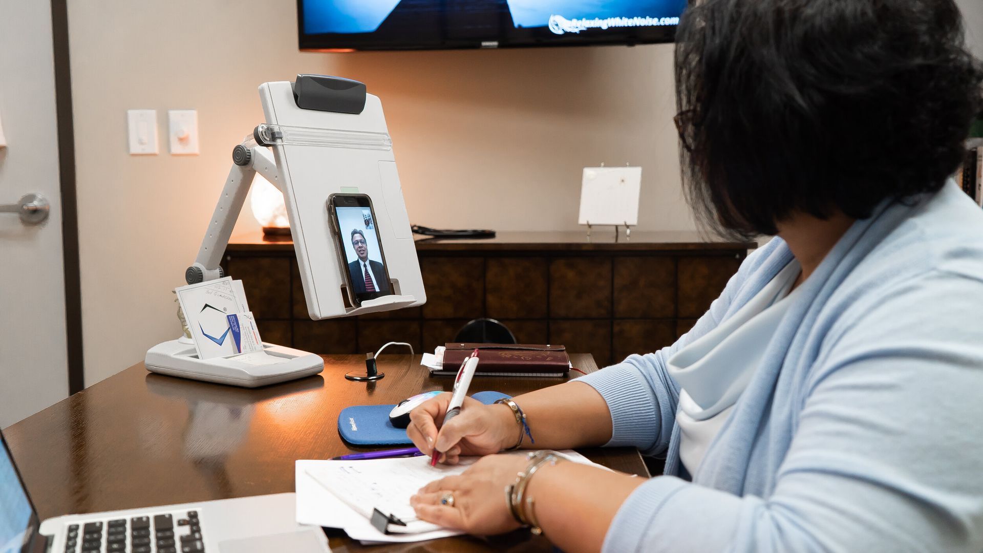 A woman is sitting at a desk with a laptop and a clipboard.
