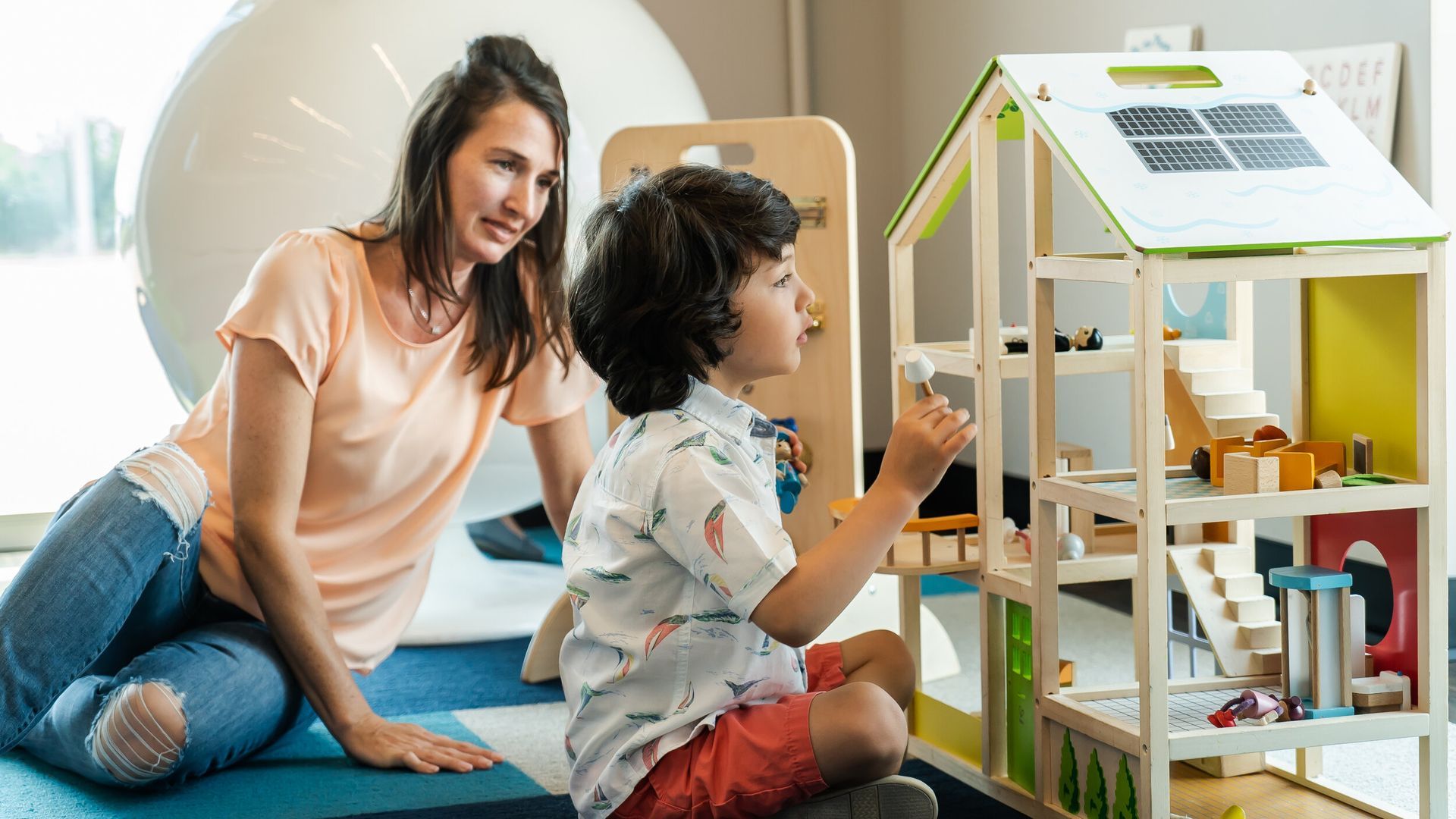 A woman and a child are sitting on the floor playing with a doll house.