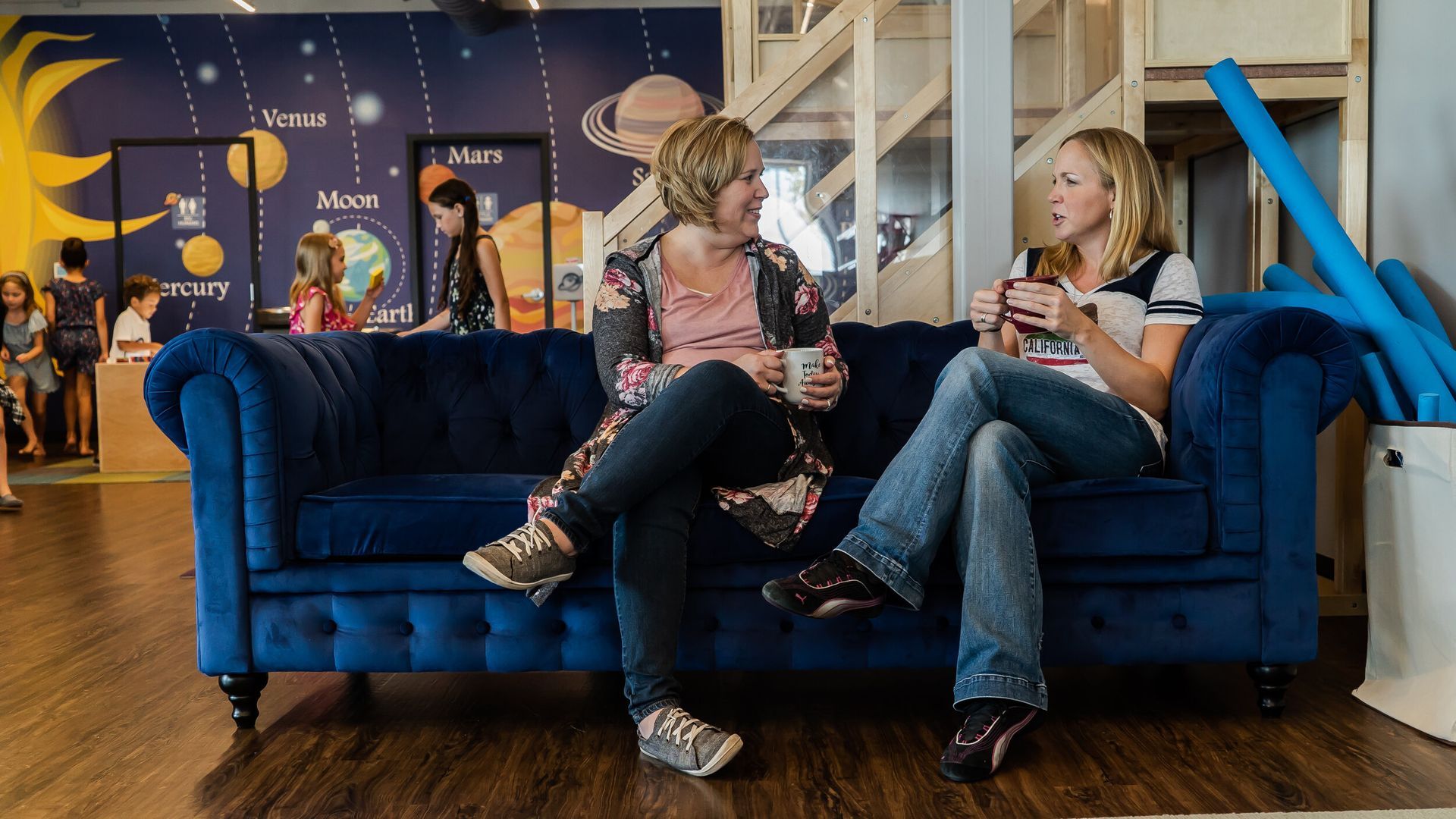 Two women are sitting on a blue couch talking to each other.