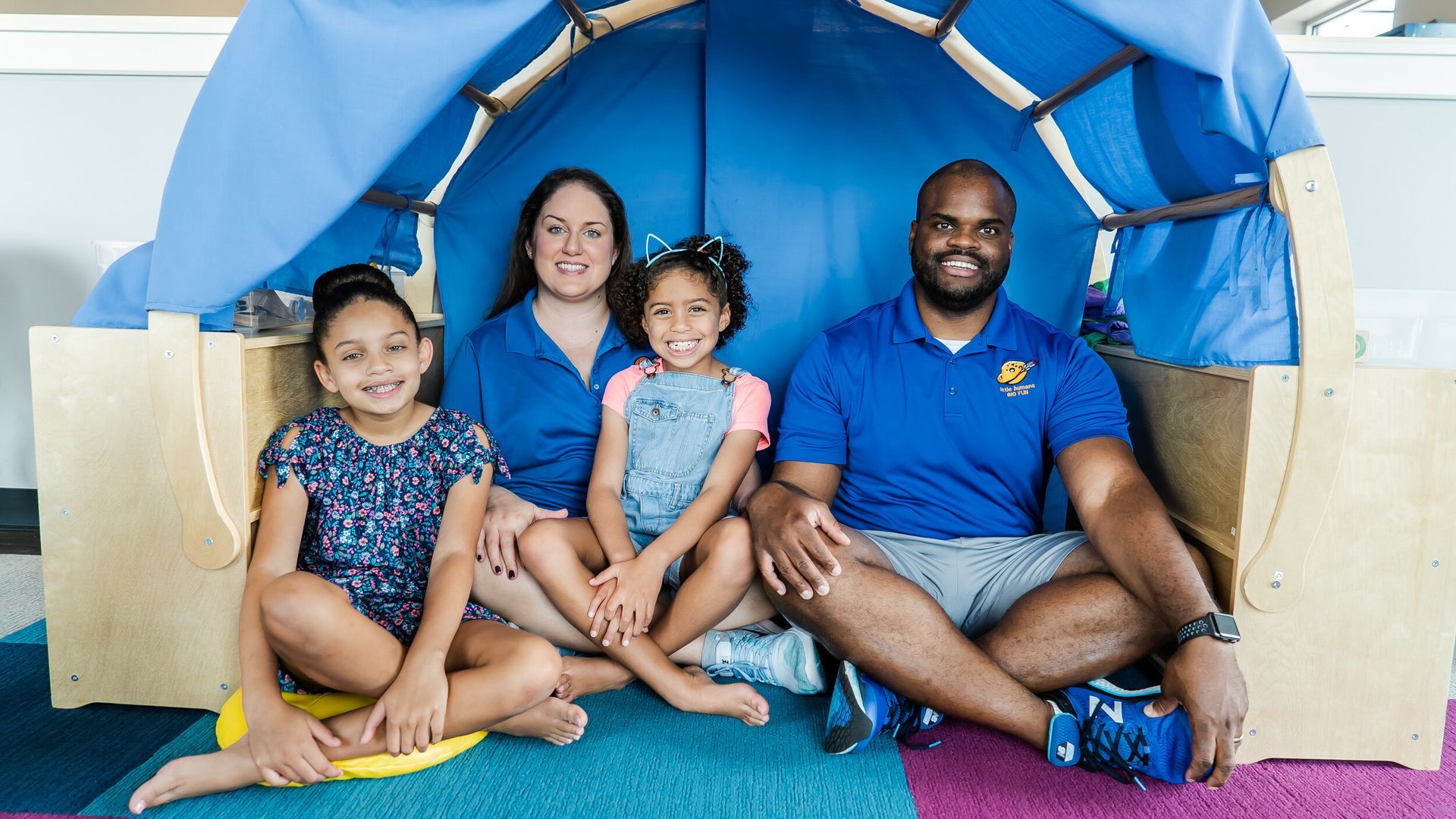 A family is sitting under a blue tent on the floor.