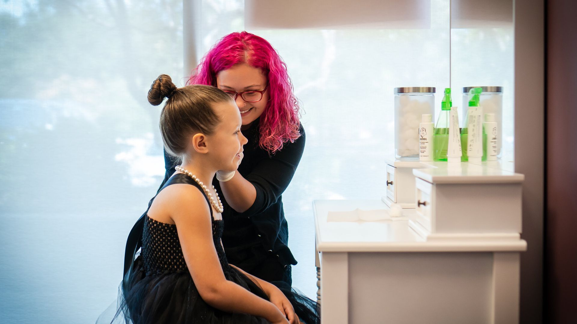 A woman with pink hair is applying makeup to a little girl.