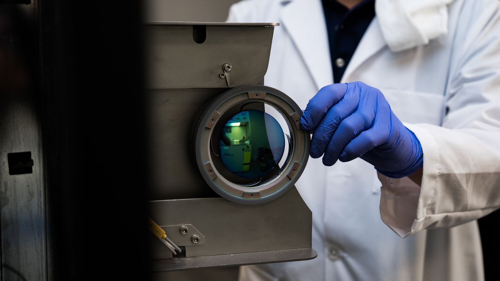 A man in a lab coat and blue gloves is looking through a lens.