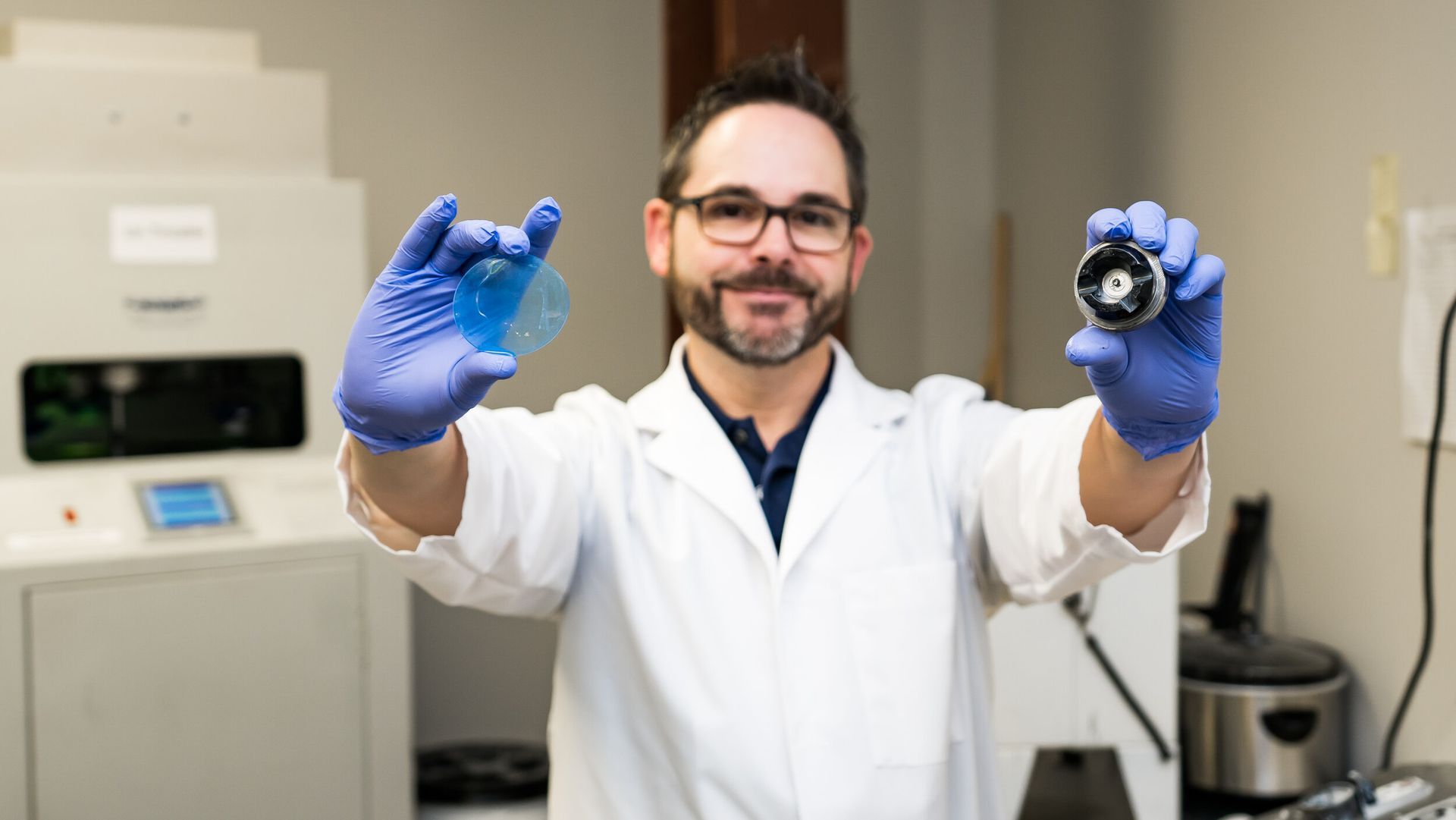 A man in a lab coat and blue gloves is holding two objects in his hands.