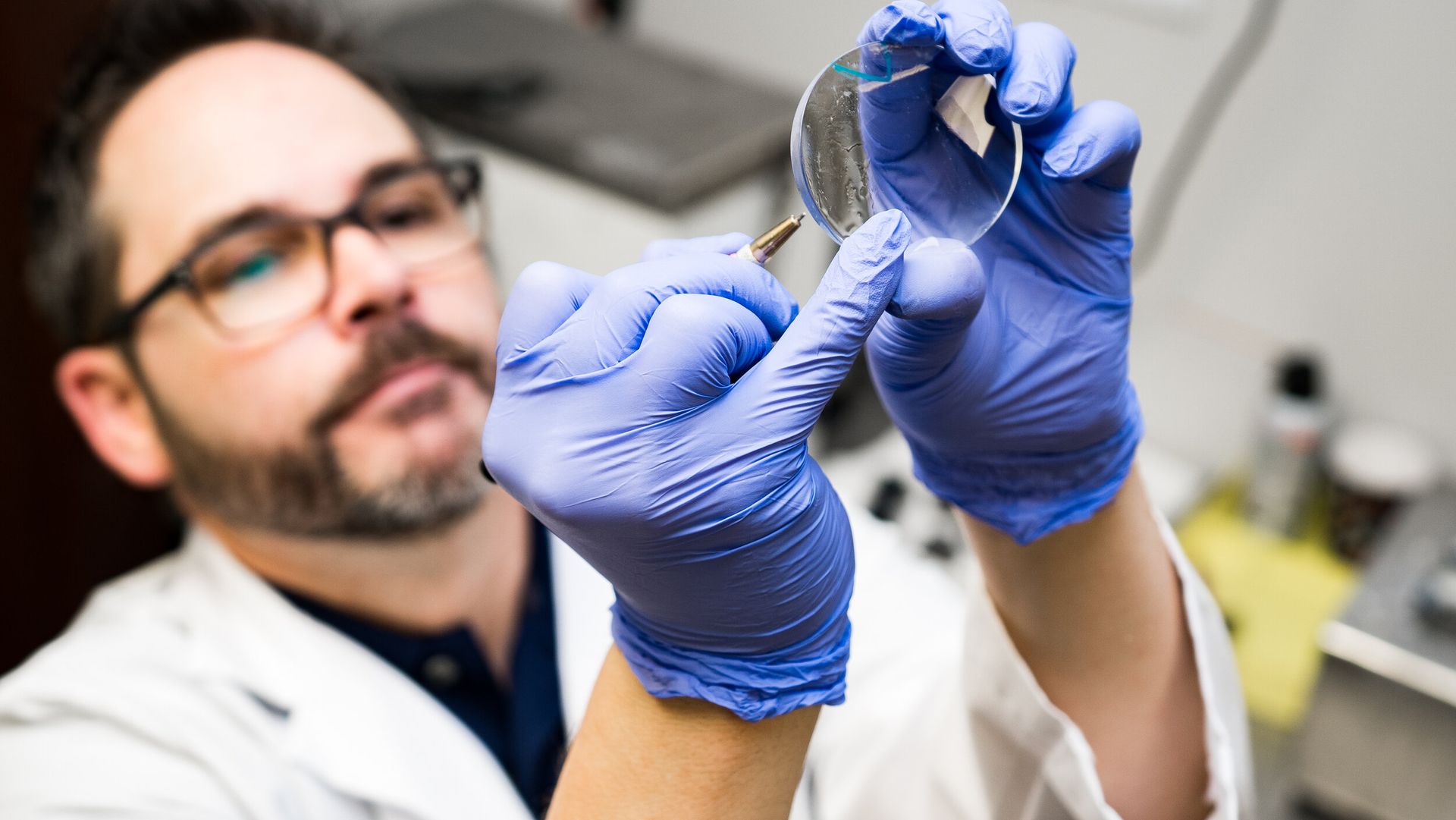 A man in a lab coat and blue gloves is holding a piece of glass.