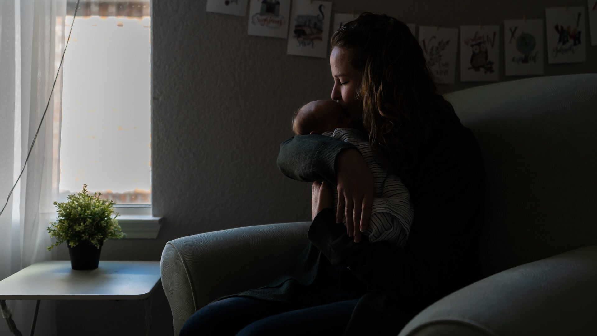A woman is holding a baby while sitting in a chair in front of a window.