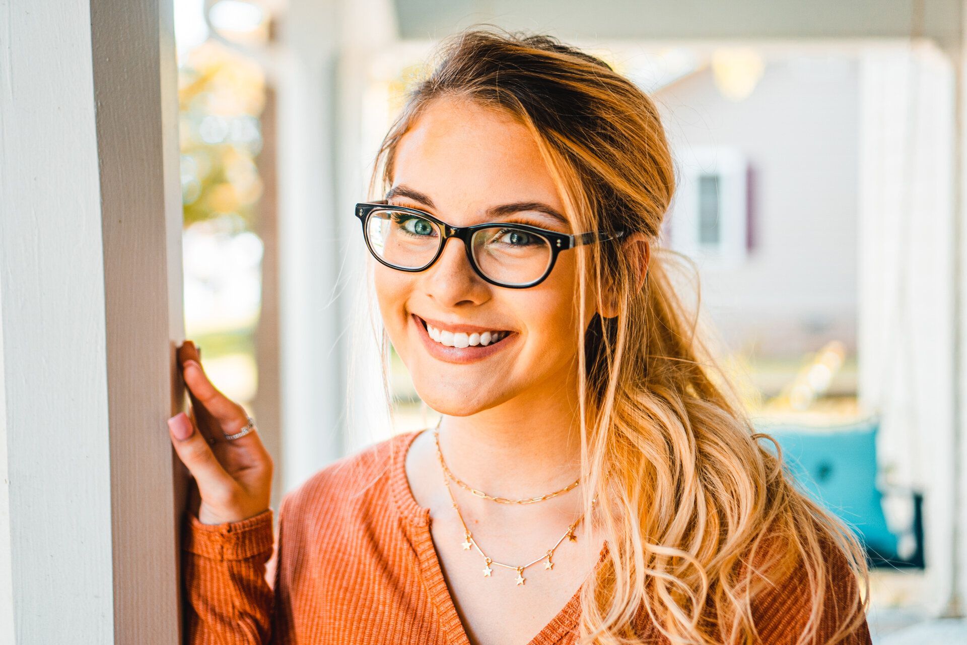 A woman wearing glasses and a necklace is smiling and leaning against a wall.