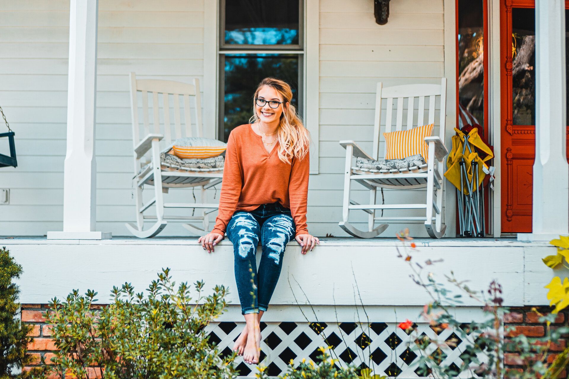 A woman is sitting on a porch next to a rocking chair.