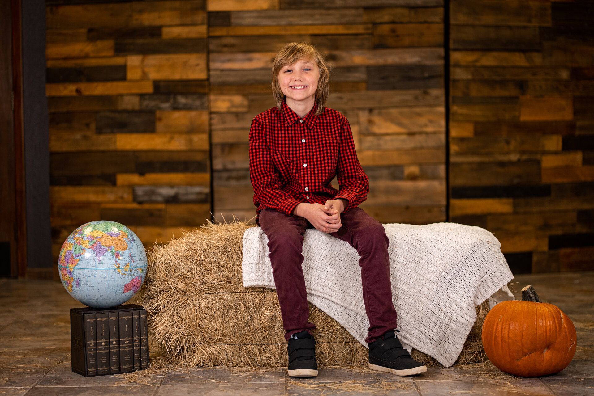 A young boy is sitting on a bale of hay next to a pumpkin and a globe.