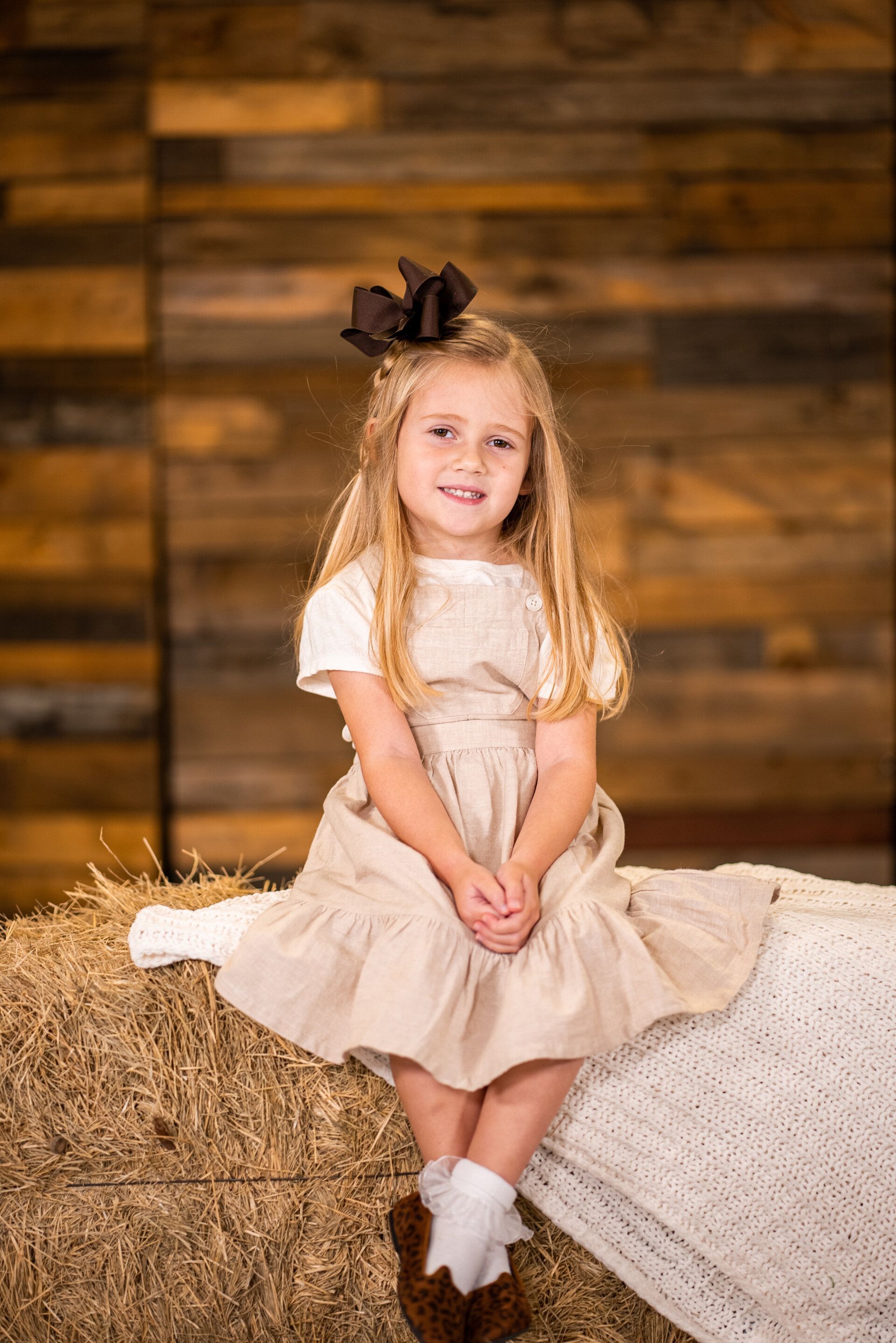 A little girl in a dress is sitting on a bale of hay.