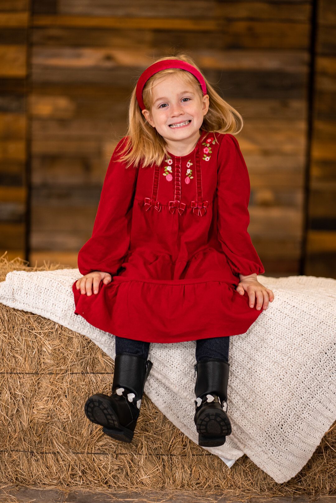 A little girl in a red dress is sitting on a bale of hay.