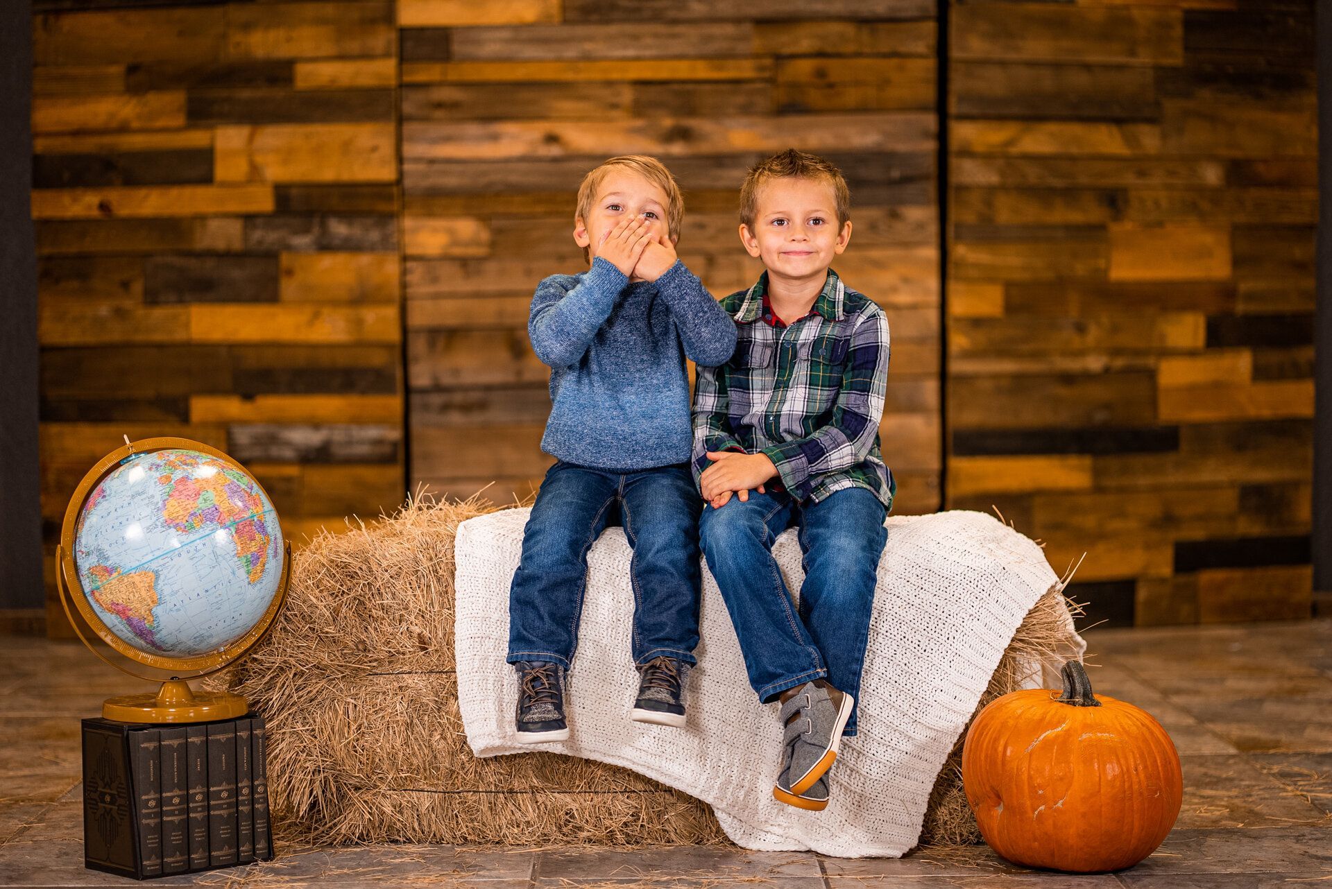 Two young boys are sitting on a bale of hay next to a pumpkin and a globe.