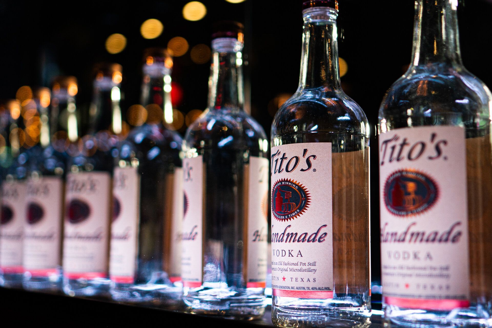 A row of tito 's vodka bottles are lined up on a bar.