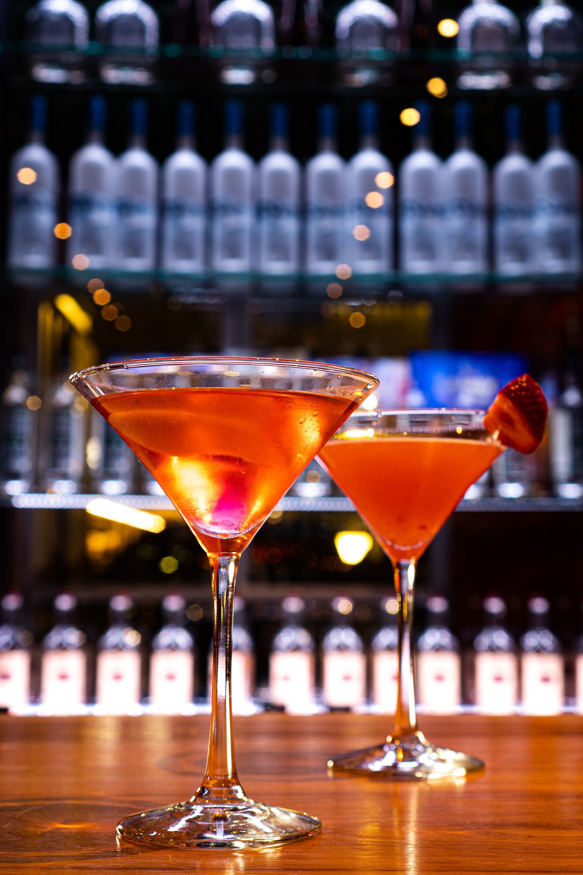 Two martini glasses on a bar with bottles in the background