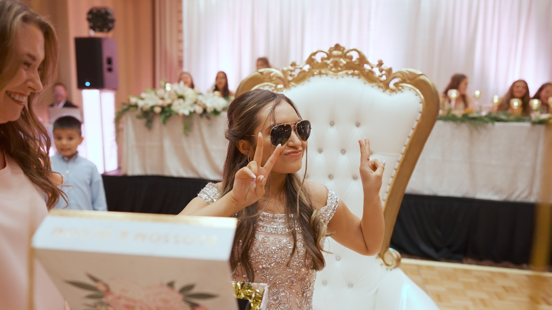 A little girl in a wedding dress is wearing sunglasses and making a peace sign.