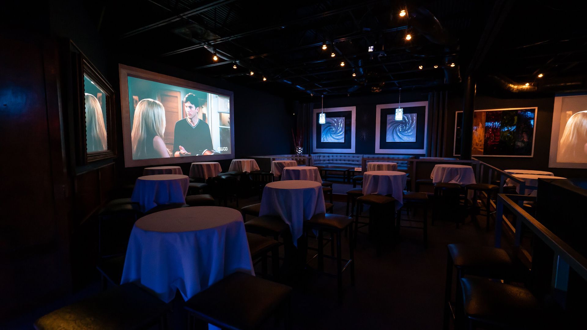 A dark room with tables and chairs and a large screen.