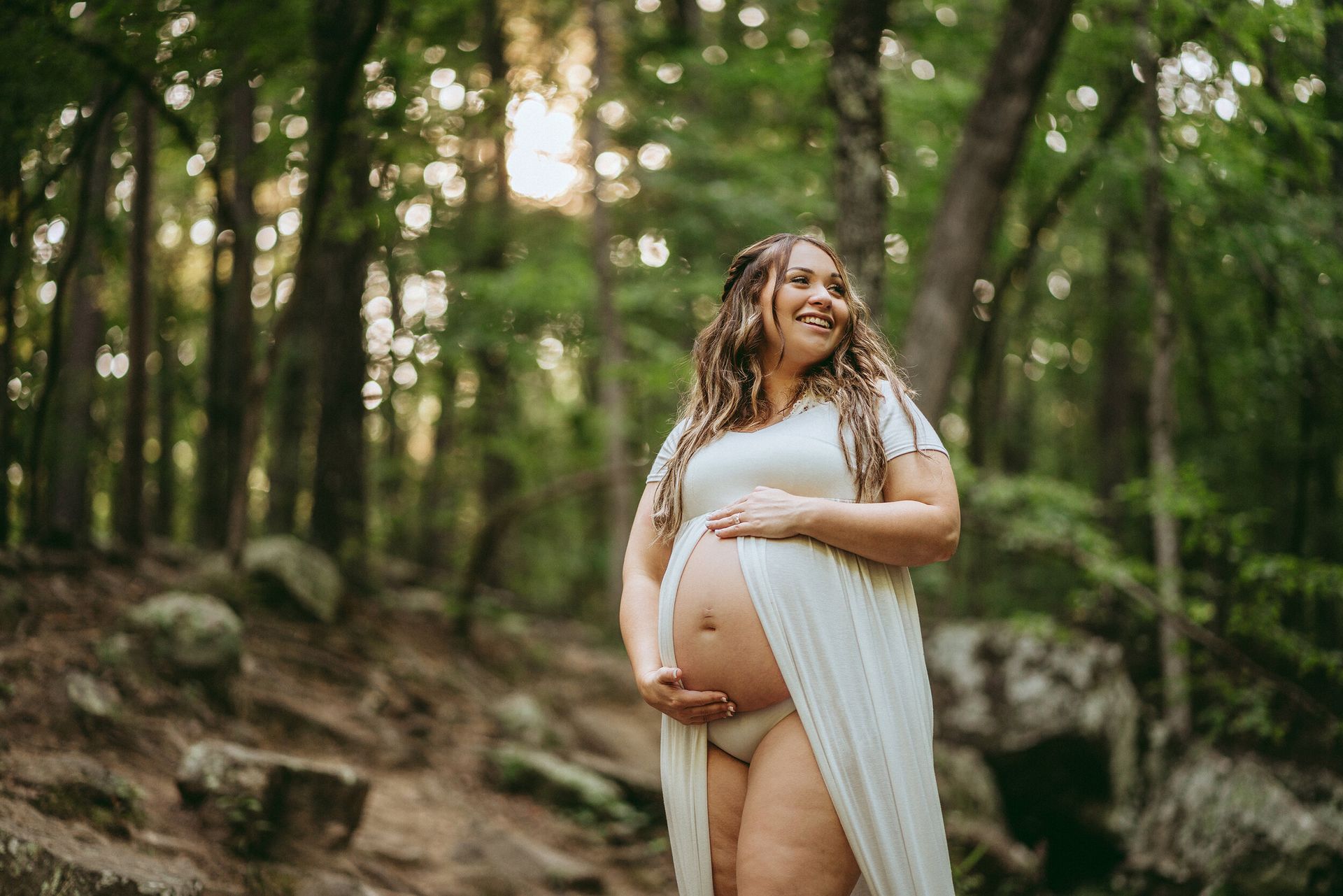 A pregnant woman in a white dress is standing in the woods holding her belly.