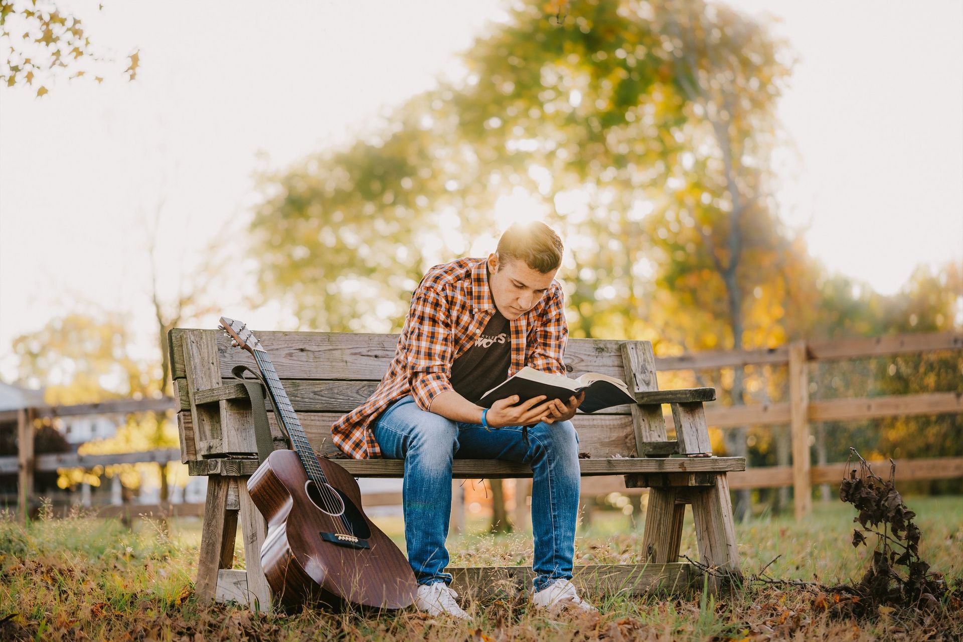 A man is sitting on a bench reading a book next to a guitar.