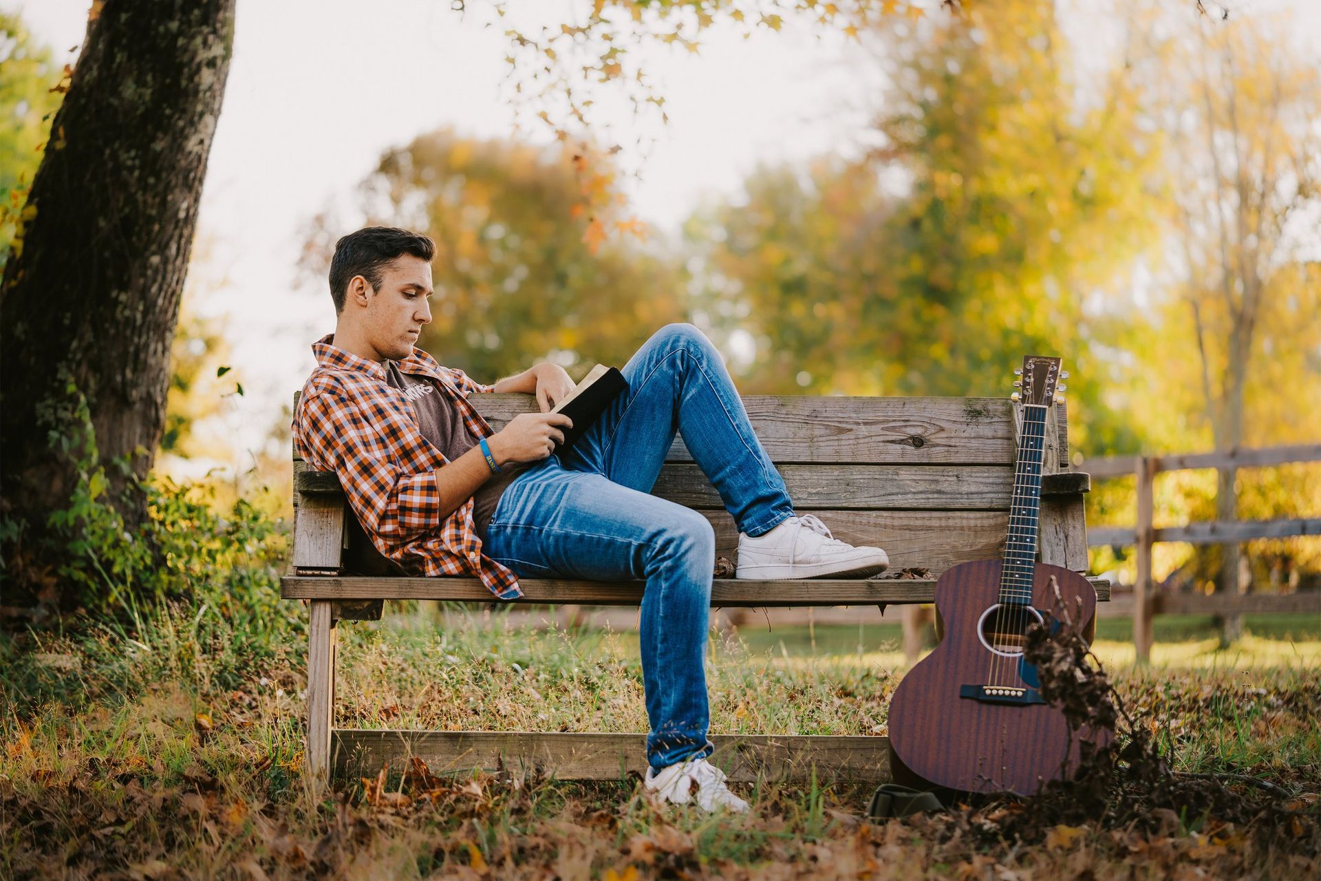 A man is sitting on a bench reading a book next to a guitar.
