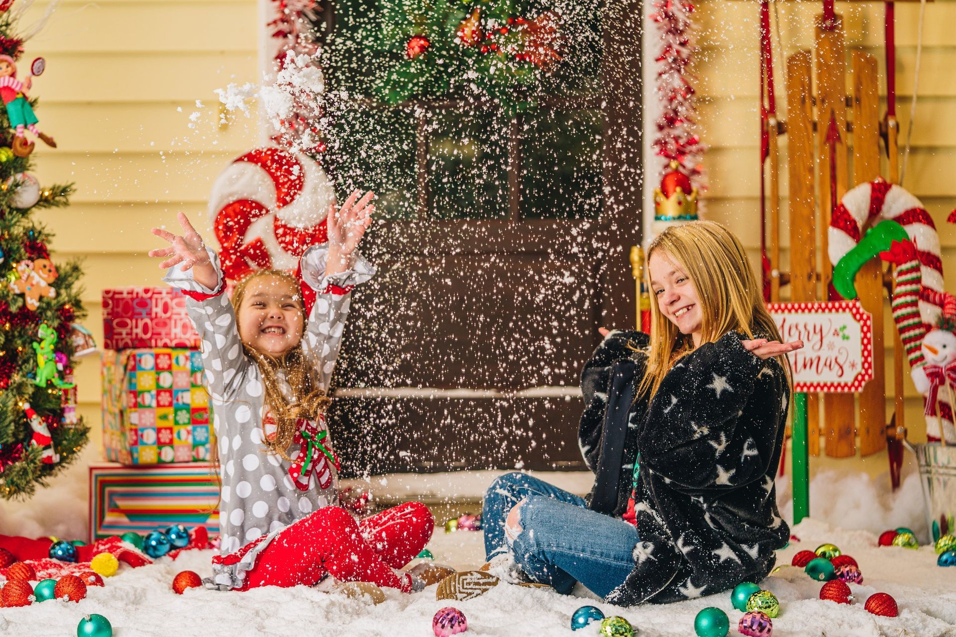 Two young girls are sitting in the snow with christmas decorations.
