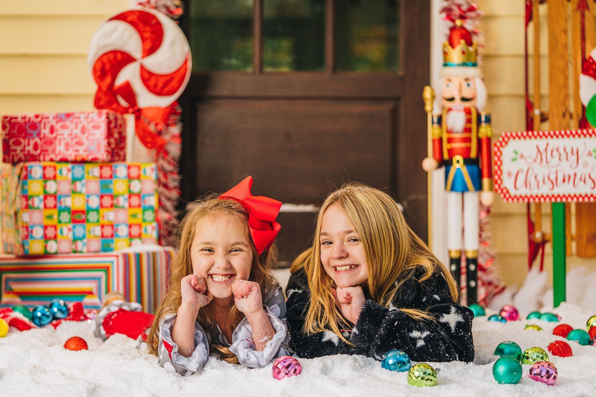 Two little girls are laying in the snow on a porch decorated for christmas.