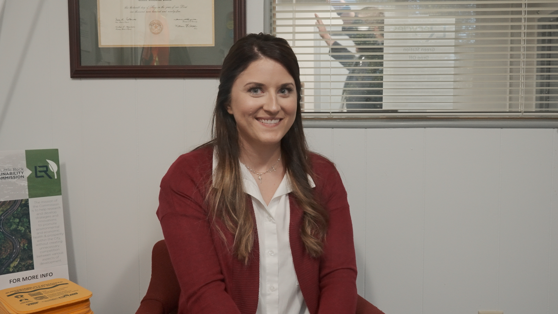 A woman in a red sweater is sitting at a desk in front of a window.