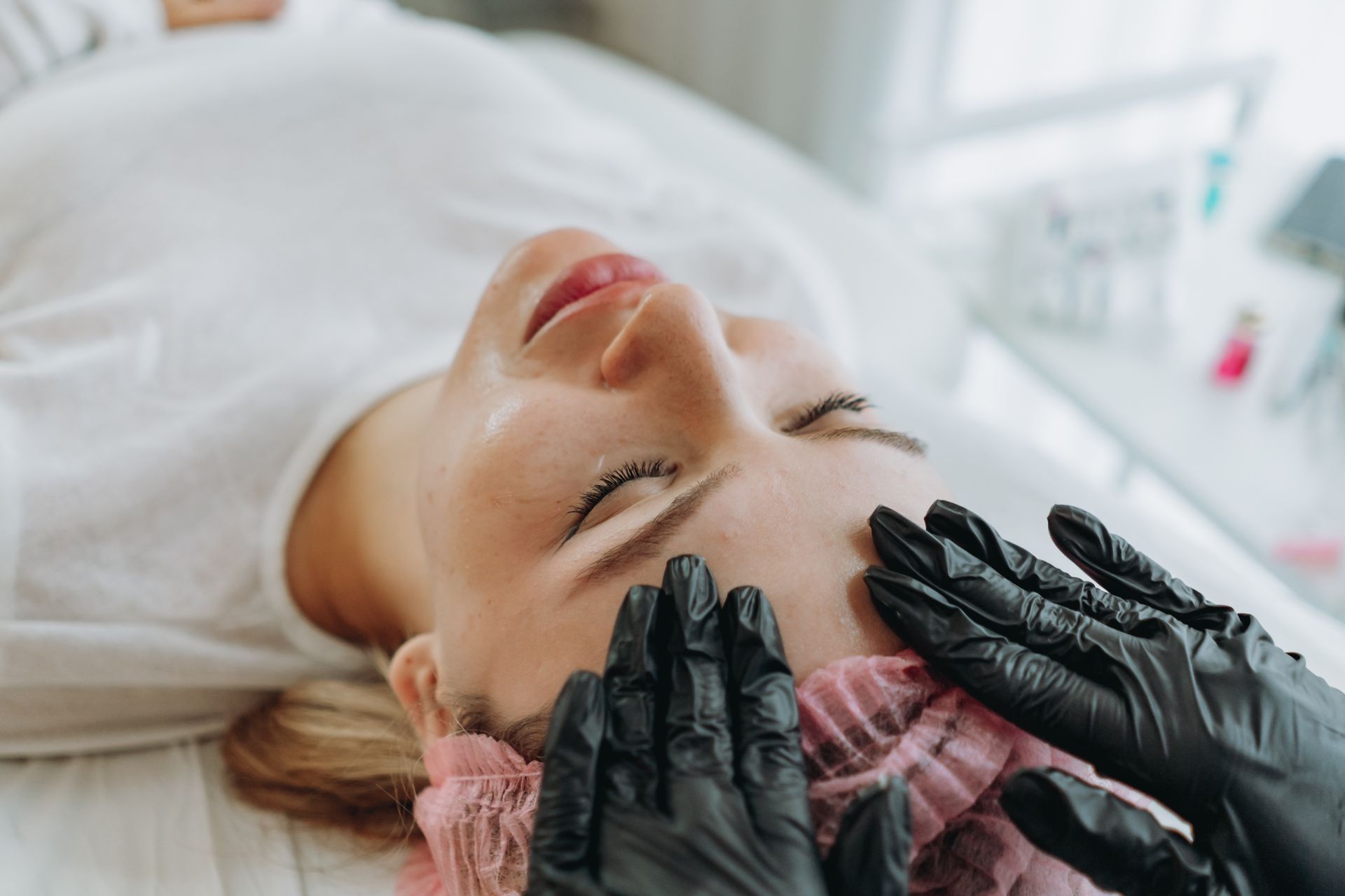 Woman receiving a facial treatment; gloved hands on her forehead, eyes closed.