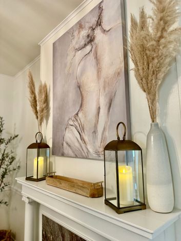 A living room with a fireplace , lanterns , pampas grass and a painting on the wall.