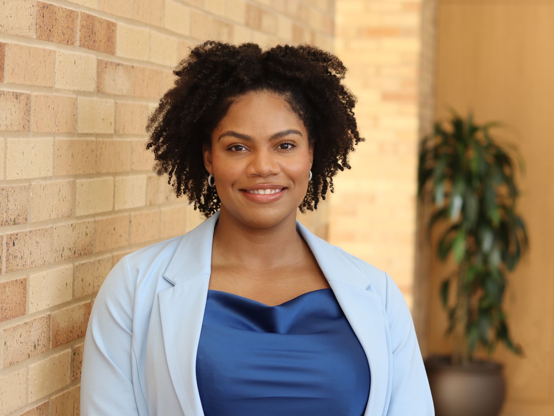Smiling woman with long, curly brown hair wearing a light purple dress against a white background.