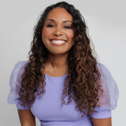 Smiling woman with long, curly brown hair wearing a light purple dress against a white background.