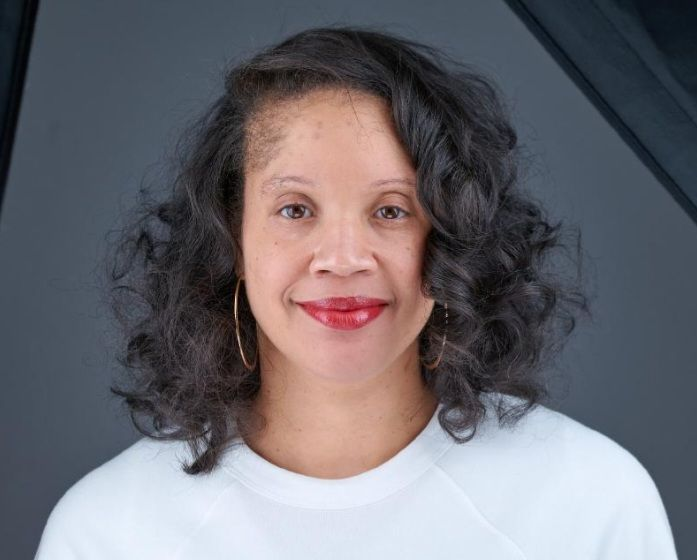 Smiling woman with long, curly brown hair wearing a light purple dress against a white background.