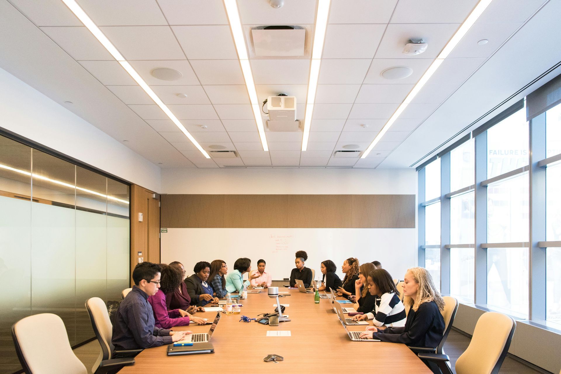 A meeting in a modern office. People are seated around a long table, laptops out, discussing.