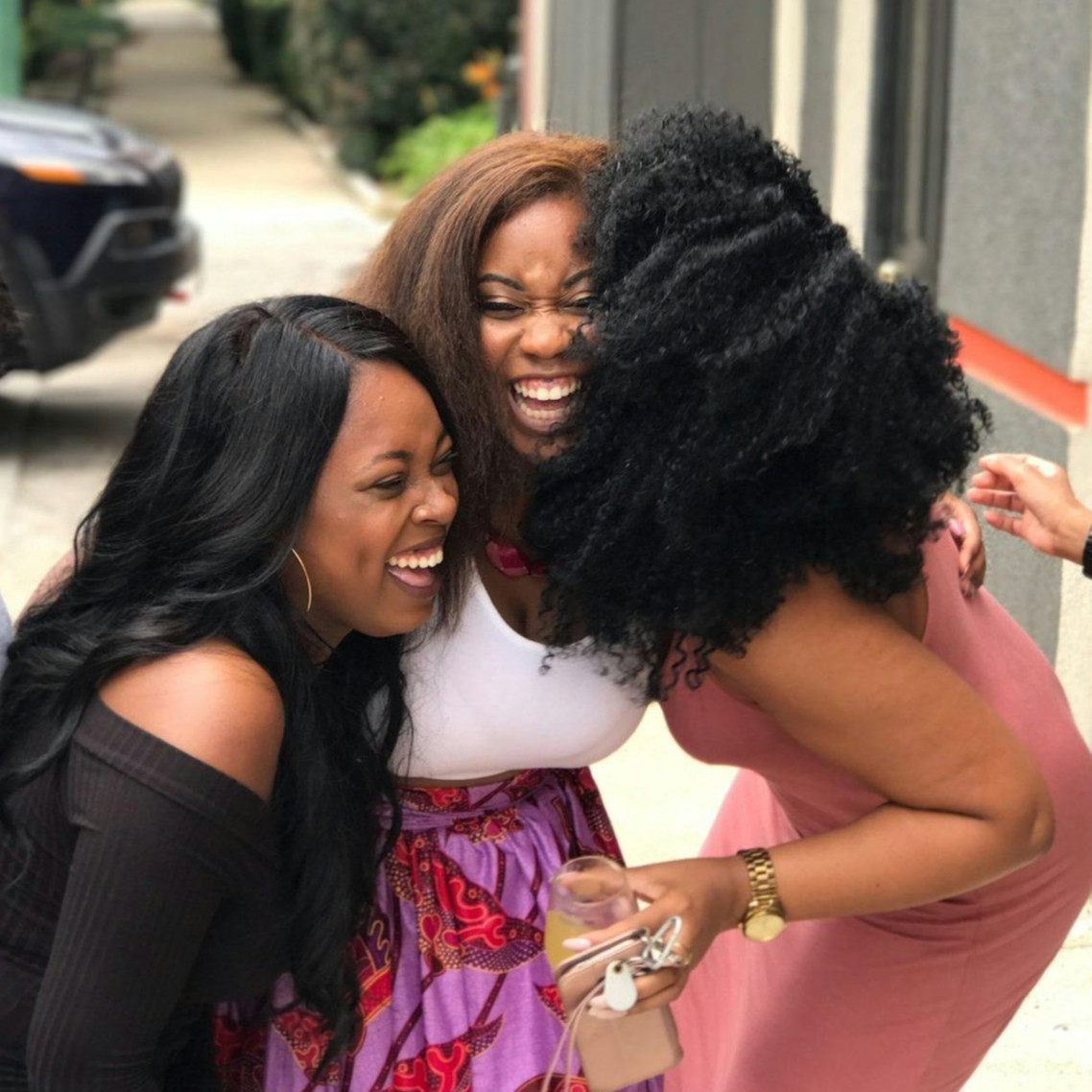 Three laughing women embracing outdoors. Two in dark clothing, one in a pink dress and African print skirt.