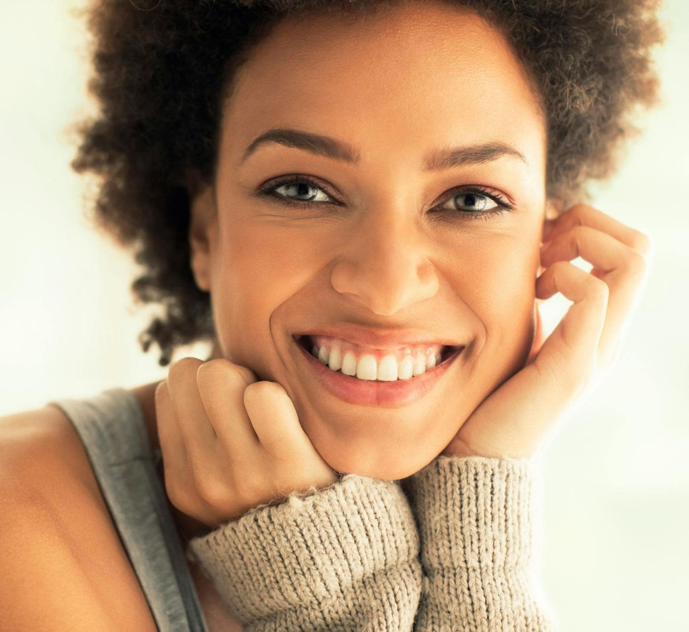 Woman with curly hair smiles, resting chin on hands, wearing a gray sweater.