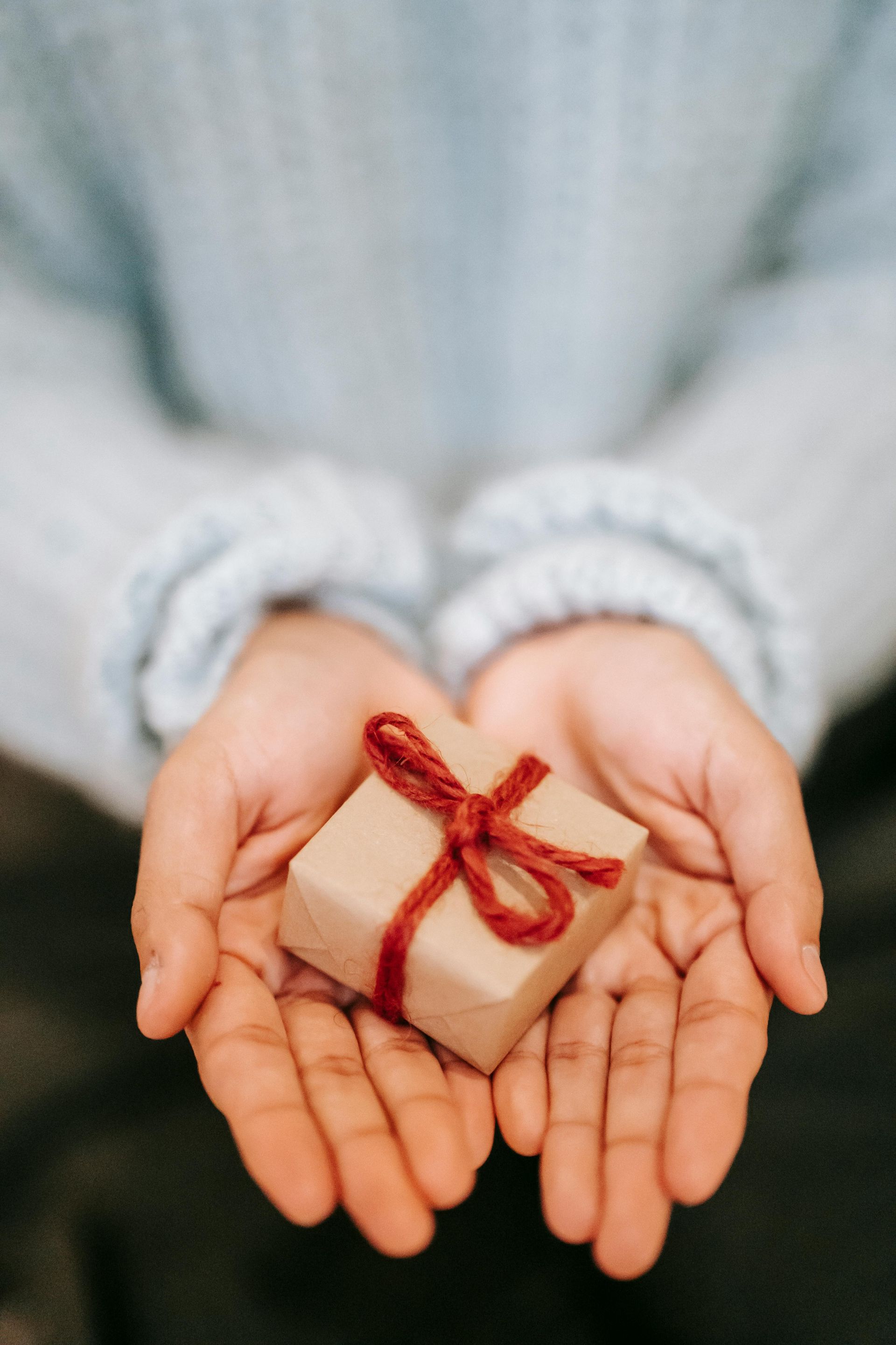 Hands cupping a small, brown gift box tied with a red string bow, against a light blue sweater.