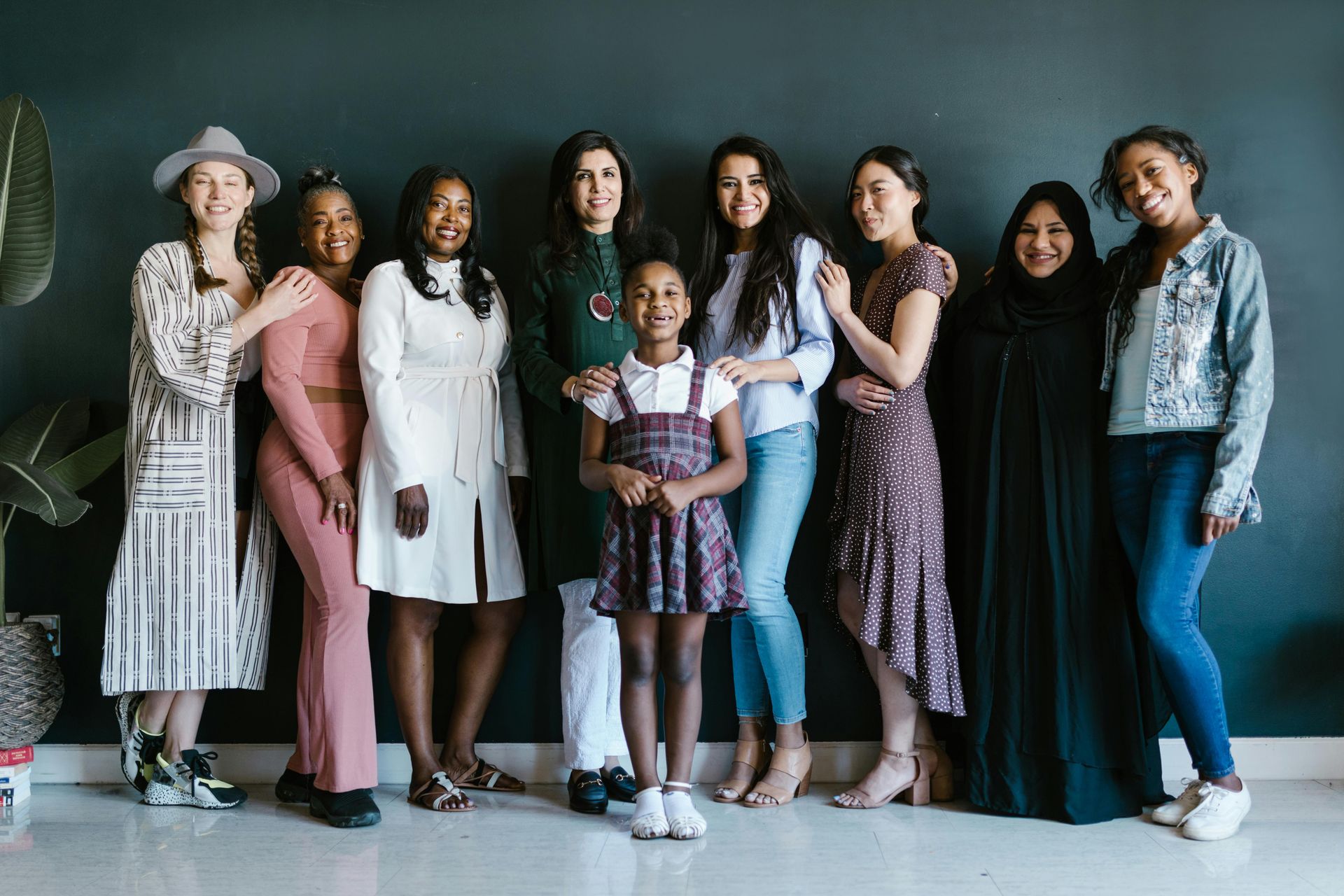 Group of diverse women and a girl, smiling and posing together against a dark wall.