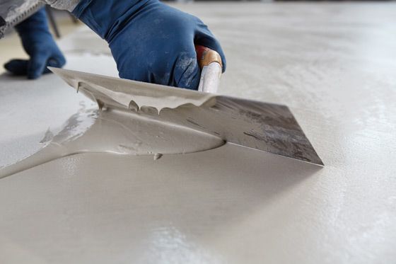 A gloved hand uses a metal trowel to smooth wet, light-gray cement over a floor surface.