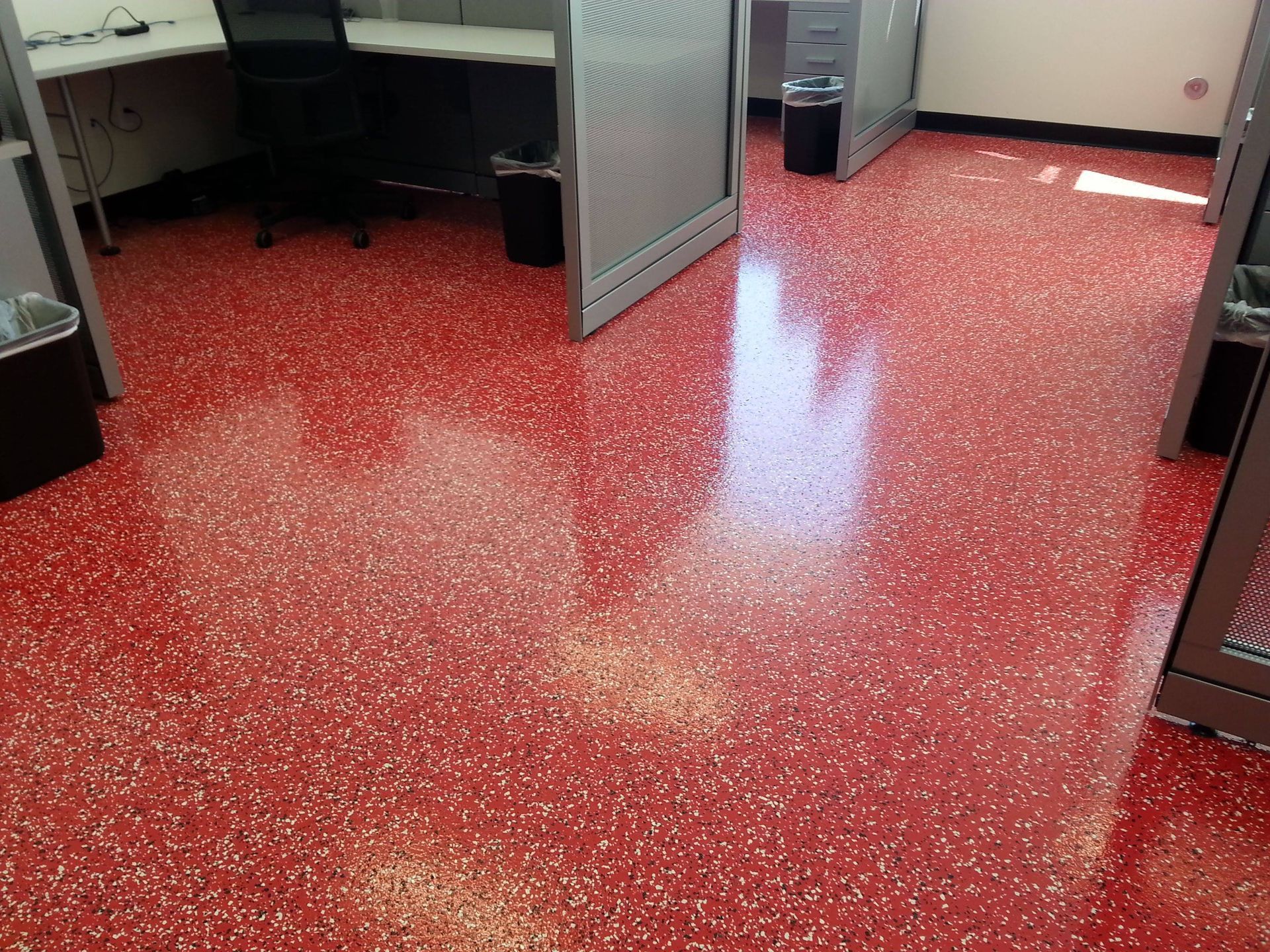 An office interior featuring a shiny red floor with white speckles, arranged with workstations and grey partitions.