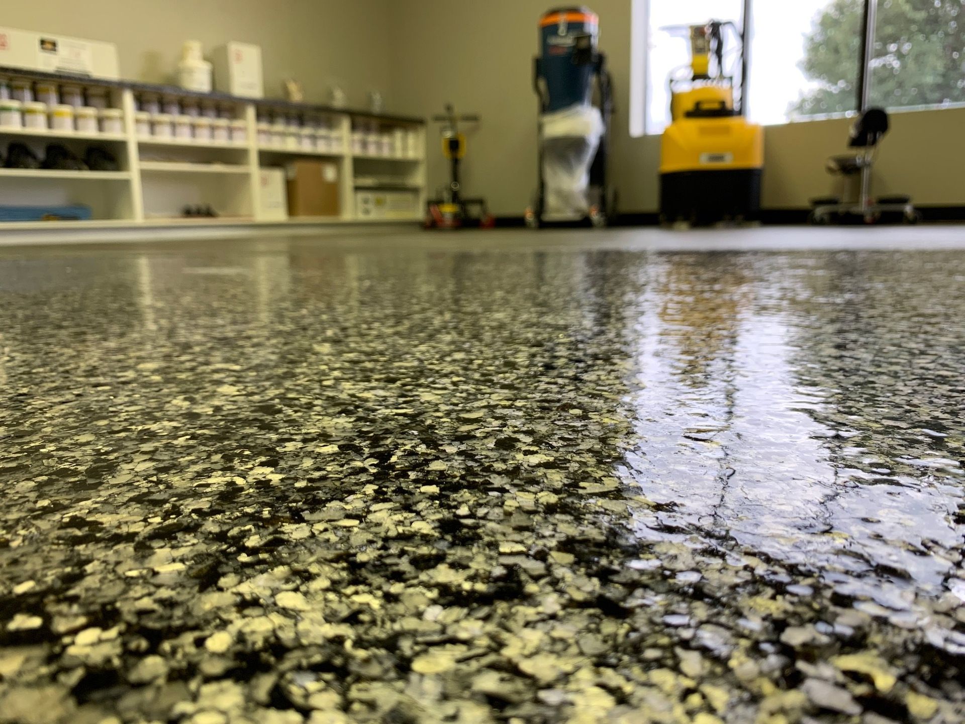 A close-up of a polished concrete floor with black and white flakes, with storage shelves and equipment in the background.