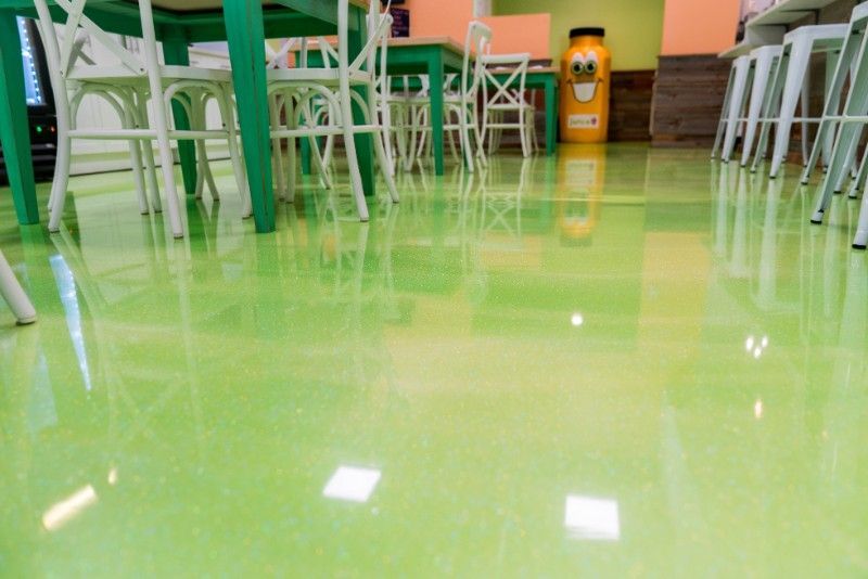 A brightly lit cafe interior featuring a polished, glossy lime-green epoxy floor, white chairs, and green tables.