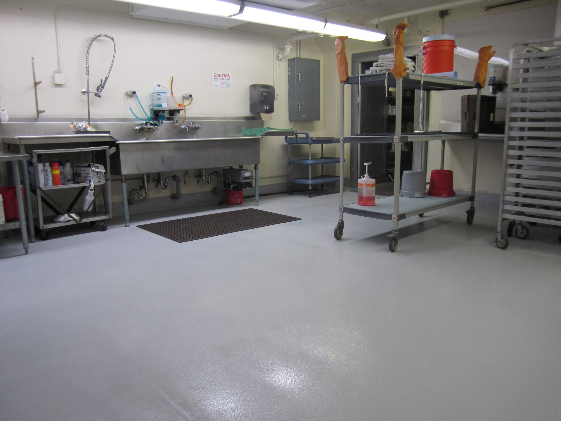 A commercial kitchen dishwashing station with stainless steel sinks, a floor mat, rolling metal carts, and storage shelves.