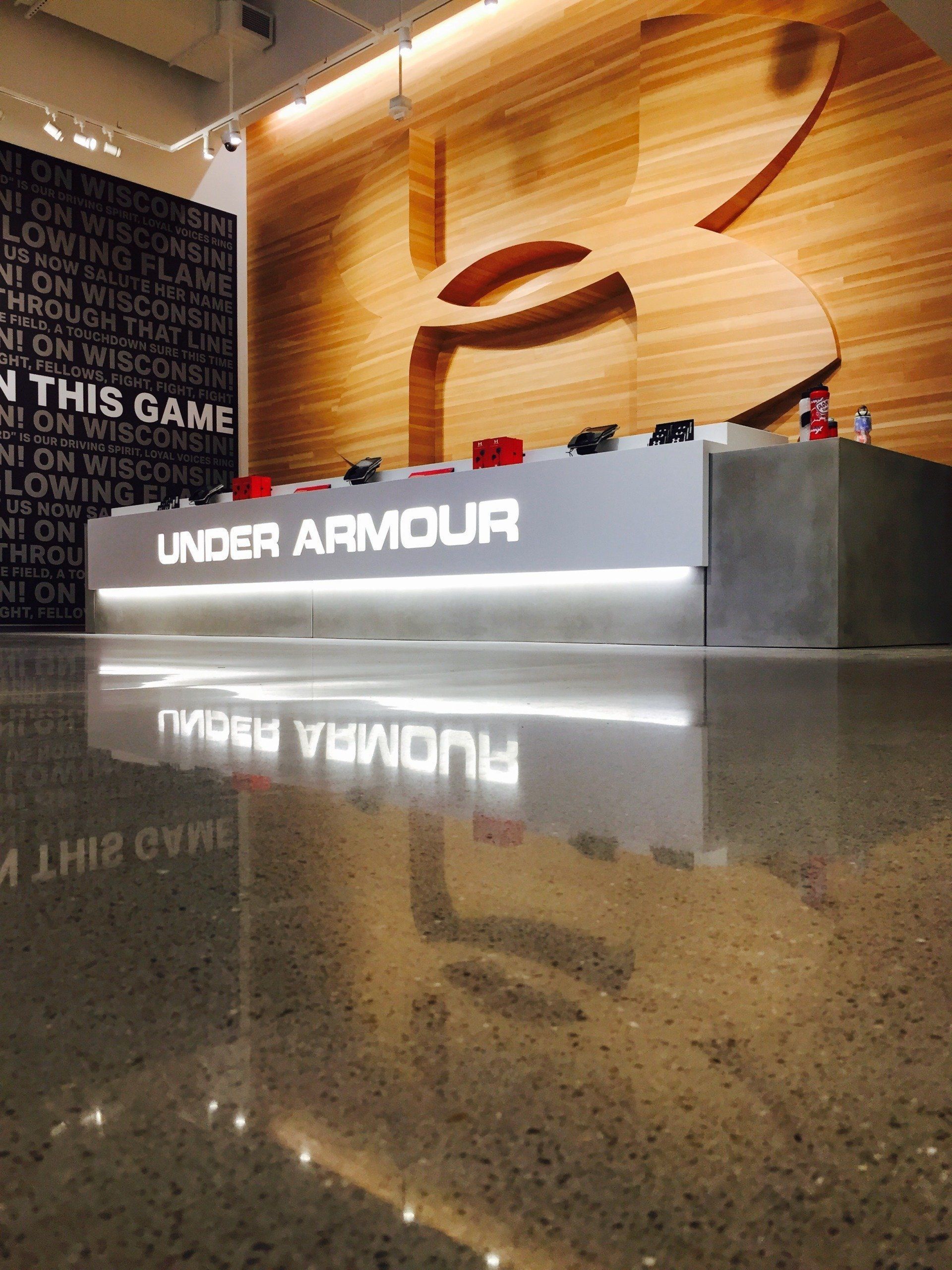 A modern Under Armour store counter with a large logo on a wood-paneled wall, reflected in a polished concrete floor.