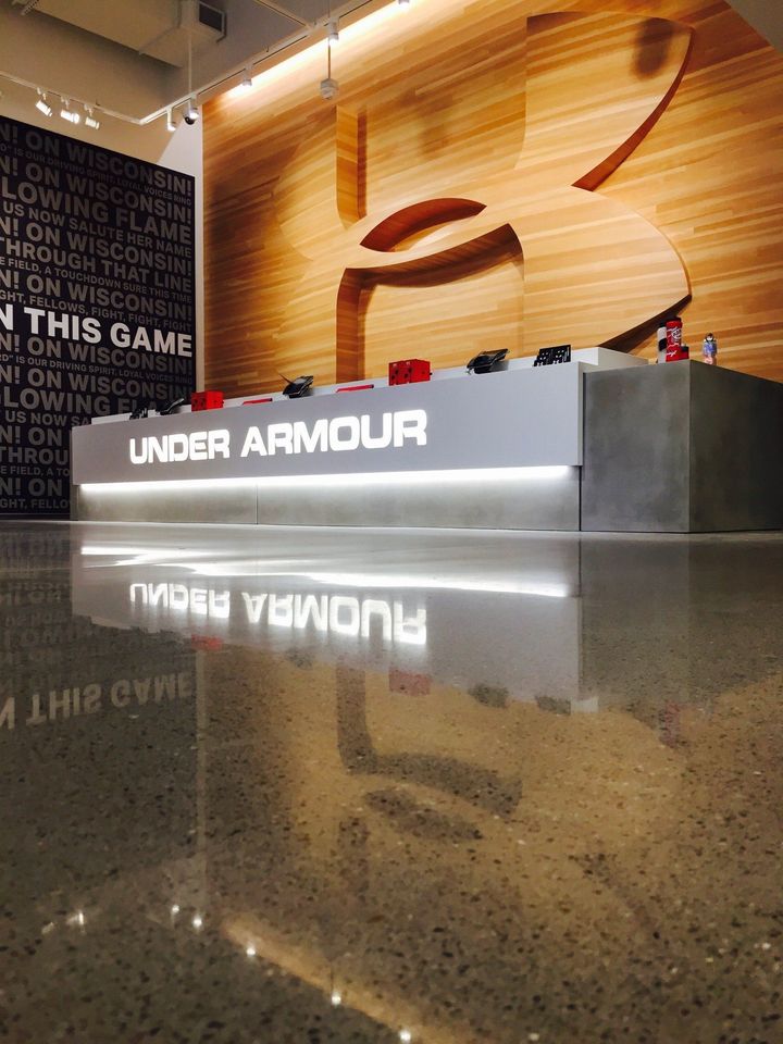 A sleek Under Armour store counter with a large logo on a wood-paneled wall, reflected in a polished concrete floor.