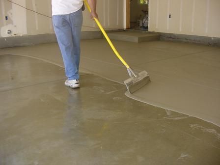 A person uses a long-handled tool to smooth wet concrete across a garage floor.