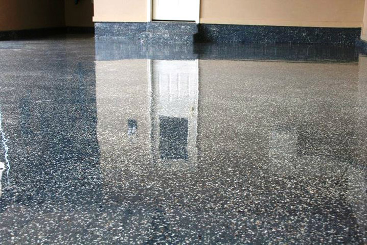 A polished, dark-colored garage floor with speckled decorative flakes, reflecting the light and an interior door.