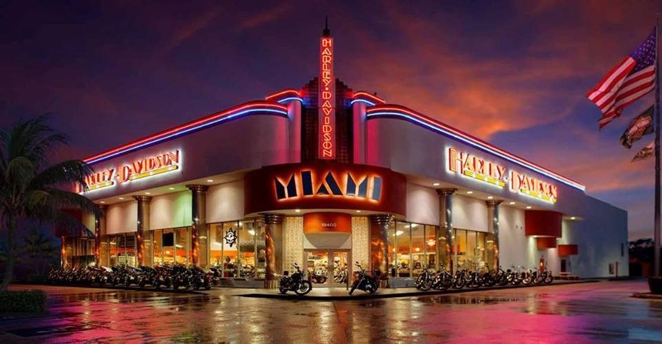 A neon-lit Harley-Davidson dealership in Miami at twilight, with parked motorcycles in front and an American flag outside.